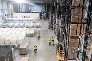 Aisle view of Basildon warehouse with high-rack pallets and staff in hi-vis vests during fire-safety inspection in an Essex logistics hub.