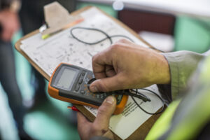 Electrician using a digital multimeter to perform PAT testing, with circuit diagrams on a clipboard in the background