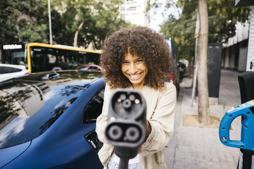 A smiling woman with curly hair holds an electric vehicle charging plug towards the camera, standing next to a blue EV at a public charging station on a city street.